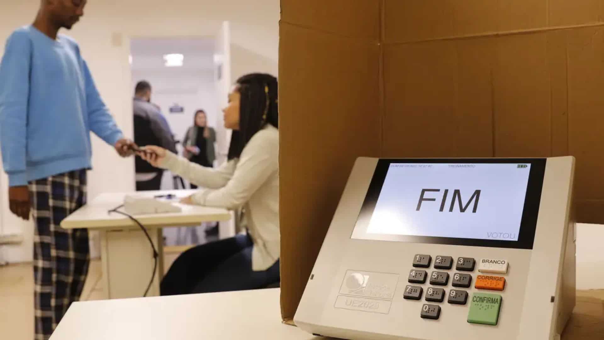 Seção de votação com urna eletrônica e pessoas ao fundo